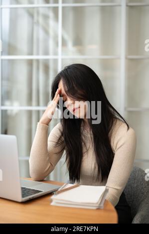 Une jeune étudiante asiatique stressée et fatiguée est assise à sa table d'étude à la maison, faisant des devoirs difficiles ou se préparant à l'examen. Banque D'Images