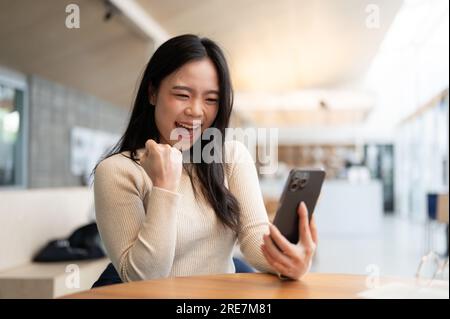 Une jeune femme asiatique joyeuse regarde l'écran de son smartphone avec un visage enjoué et se réjouit après avoir reçu de bonnes nouvelles tout en étant assise dans un caf Banque D'Images