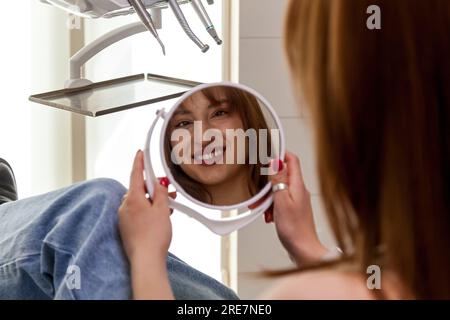 Une belle fille sourit à son reflet dans le miroir. Une fille avec de belles dents blanches sourit après un traitement chez le dentiste. Banque D'Images