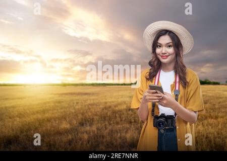 Femme asiatique avec un chapeau et un appareil photo tenant un téléphone portable voyageant sur la prairie avec un fond de scène de coucher de soleil. Concept de journée mondiale du tourisme Banque D'Images