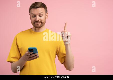 Homme satisfait levant le doigt vers le haut ont l'idée créative look sur téléphone portable isolé sur blanc studio rose Banque D'Images