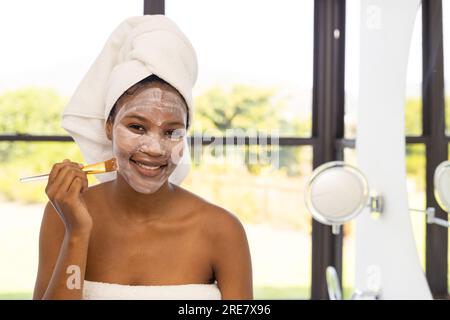 Portrait de femme afro-américaine heureuse portant une serviette sur la tête appliquant un masque facial de beauté dans la salle de bain Banque D'Images