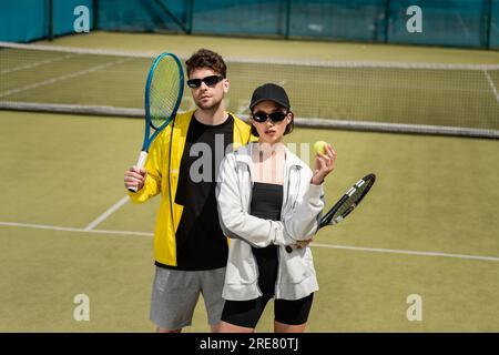 mode et sport, homme en lunettes de soleil et femme en casquette tenant raquettes et balle sur le court de tennis Banque D'Images