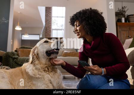 Heureuse femme biracial caressant le chien et utilisant smartphone à la maison Banque D'Images