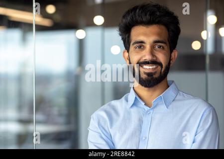 Portrait d'un jeune étudiant indien debout dans un bureau, un campus, une bibliothèque portant une chemise bleue et souriant à la caméra. Gros plan. Banque D'Images