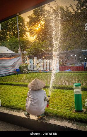 Une jardinière vietnamienne portant un chapeau conique s'accroupie alors qu'elle pulvérise de l'eau sur des parterres de fleurs dans le parc de la réunification, à Hanoi, au Vietnam. Banque D'Images