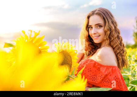 femme heureuse dans une robe rouge au coucher du soleil dans un champ de tournesols, souriant doucement Banque D'Images