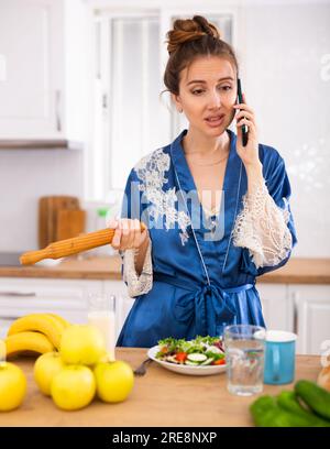 Femme insatisfaite avec la goupille dans les mains parlant au téléphone dans kichen Banque D'Images