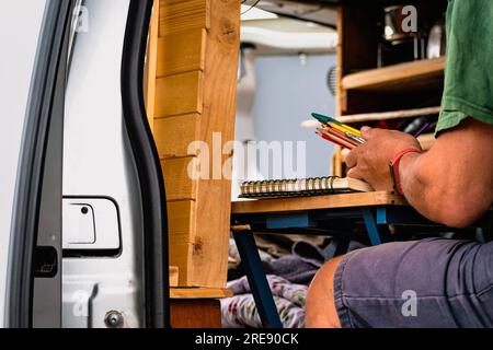 Homme dessinant sur un cahier et tenant des crayons sur une table dans un van converti Banque D'Images