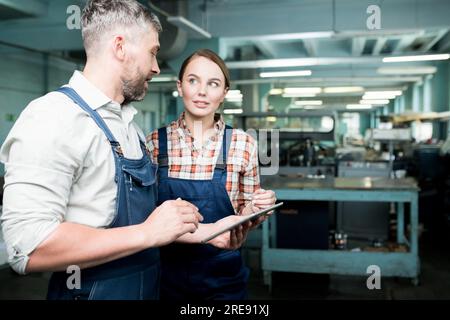 Travailleurs industriels modernes confiants dans des combinaisons debout dans l'atelier d'usine avec des machines en arrière-plan et utilisant la tablette numérique pour vérifier les données en ligne Banque D'Images