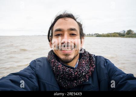 portrait selfie de jeune homme vénézuélien avec moustache et barbe, piercing nez et portant une veste bleue et une écharpe, il rit debout sur la tarte Banque D'Images