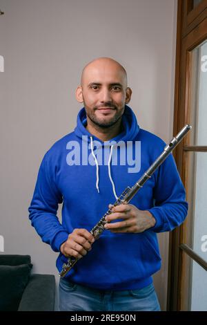 portrait vertical d'un jeune homme latino avec barbe et chauve, vêtu de bleu, heureux avec sa flûte debout à la maison regardant la caméra. Banque D'Images