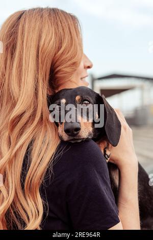 Vue latérale de la femme rousse embrassant le chien Teckel tout en marchant dans la rue ensemble pendant le week-end Banque D'Images