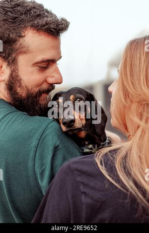 Vue latérale de l'homme souriant et la femme rousse embrassant le chien Teckel tout en marchant dans la rue ensemble pendant le week-end Banque D'Images