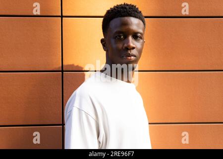 Portrait d'un homme afro-américain sérieux en t-shirt blanc regardant la caméra tout en se tenant debout contre un mur orange Banque D'Images