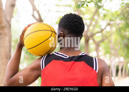 Vue arrière d'un sportif masculin afro-américain tenant le basket-ball sur l'épaule dans le parc contre des arbres brouillés pendant la journée Banque D'Images