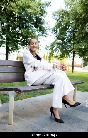 Corps entier de souriant jeune femme afro-américaine avec les cheveux longs en costume blanc et chaussures à talons hauts assis sur un banc en bois avec les jambes croisées tandis que Banque D'Images