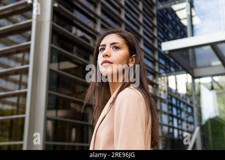 De ci-dessous vue latérale de pensive jeune femme d'affaires du Moyen-Orient dans des vêtements formels debout près du bâtiment de verre urbain et regardant loin Banque D'Images