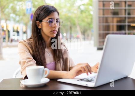 Concentré jeune femme d'affaires du Moyen-Orient réfléchi assis à table avec tasse de café et travaillant via netbook Banque D'Images