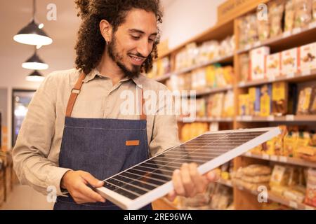 Vendeur positif mâle dans des vêtements décontractés et tablier souriant tout en se tenant près des étagères tenant et regardant le panneau solaire portable dans un supermarché moderne Banque D'Images