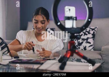 Influenceur féminin positif assis à la table avec smartphone sur trépied et lampe annulaire enregistrant vlog de beauté tout en nettoyant la brosse de maquillage avec de l'eau Banque D'Images