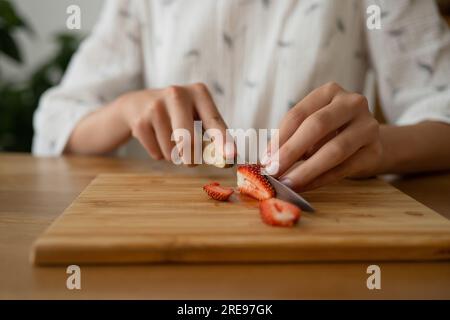 Coupez la femelle anonyme dans le tissu blanc assis et coupant la fraise rouge sur le bord au-dessus de la table en bois tout en faisant de la confiture de fraise à l'intérieur Banque D'Images
