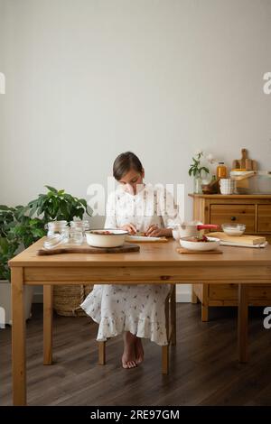 Corps complet de jeune femme en robe assise à la table en bois avec bol de fraises et bouteilles en verre tout en faisant de la confiture de fraises dans la chambre Banque D'Images