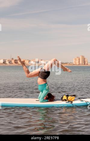 Vue latérale de la femme flexible en vêtements de sport faisant la pose de yoga avec la tête et les jambes écartées tout en pratiquant le yoga sur planche de SUP en mer sous le ciel bleu Banque D'Images