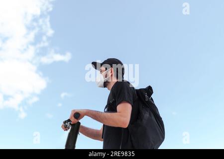 Vue latérale à angle bas d'un homme anonyme en masque respiratoire et des vêtements noirs conduisant le scooter électrique sous un ciel bleu nuageux tout en regardant vers l'avenir Banque D'Images