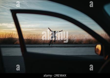 De la vue de la voiture de tout le corps jeune homme dans une ambiance décontractée portez des sauts sur une planche à roulettes tout en effectuant un kickflip sur une route en asphalte contre le ciel sombre Banque D'Images