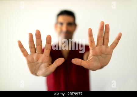 Thérapeute masculin adulte hispanique souriant flou montrant les mains avec les doigts ouverts tout en se tenant dans le salon près du mur blanc Banque D'Images