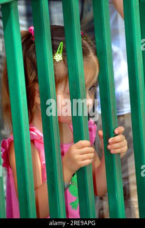 Une petite fille fait un peeks à travers des barres de fer vertes. Piégée à l'intérieur, elle est solennelle et souhaitant qu'elle soit à l'extérieur. Banque D'Images