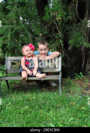 Frère et sœur posent dehors sur le banc du parc. Les enfants sont entourés d'herbe verte et d'arbres. Banque D'Images