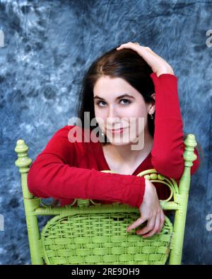 Reposant sur une chaise tournée vers l'arrière, cette adolescente sourit avec ses yeux alors qu'elle se penche à l'arrière d'une chaise en bois vert. Banque D'Images