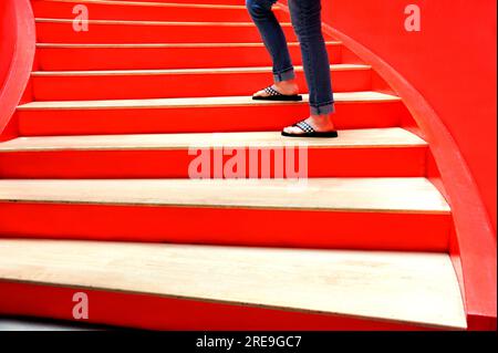 Escalier rouge monte vers le ciel. Jambes habillées en jean et tongs à carreaux noirs s'interrompent lors de sa montée. Banque D'Images