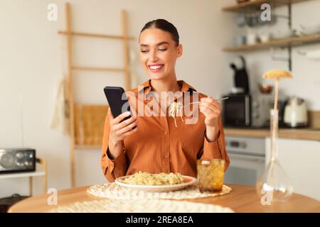 Addiction aux gadgets. Heureuse jeune femme caucasienne déjeunant dans la cuisine à la maison, mangeant des pâtes et utilisant le téléphone portable Banque D'Images