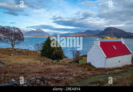 Petit cottage au toit en étain rouge au bord d'un loch surplombant les montagnes à Torridon, en Écosse Banque D'Images