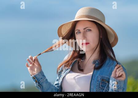 Mystérieuse femme adulte avec les yeux gris regardant la caméra. Hipster habillé en chapeau de paille, veste en denim. Femme debout sur fond naturel flou Banque D'Images