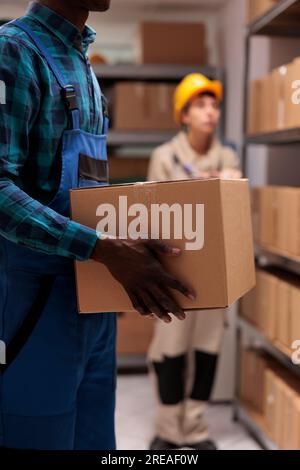 Homme afro-américain tenant le colis du client dans l'entrepôt de centre de tri de courrier. Assistant de stockage du service de livraison portant une boîte en carton et se tenant près de l'étagère avec les colis Banque D'Images