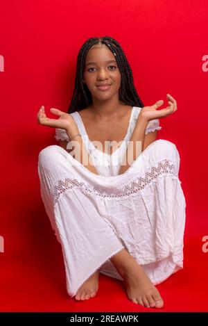 Belle jeune femme noire assise souriante à l'appareil photo. Isolé sur fond rouge Banque D'Images