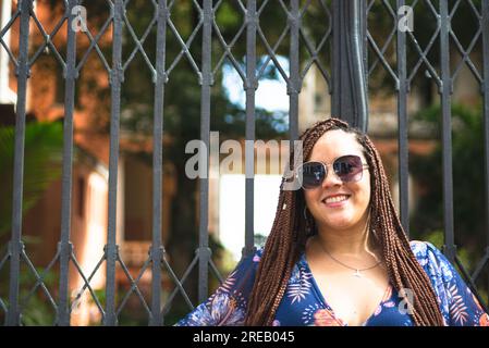 Magnifique femme avec des tresses dans ses cheveux et portant des lunettes de soleil appuyées contre une rampe de fer. Pelourinho, Brésil. Banque D'Images