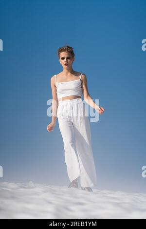 Femme blonde à la mode marche à travers la neige avec un brouillard léger sur une journée ensoleillée avec un ciel bleu. Authenticité adulte femme habillée en haut court blanc, pantalon Banque D'Images