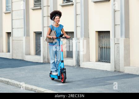 Corps complet de jeune dame noire joyeuse avec coiffure afro chevauchant scooter électrique sur la rue de la ville dans la lumière du soleil Banque D'Images