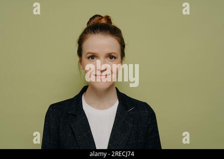 Heureuse jeune femme dans des vêtements décontractés, cheveux en chignon, souriant avec confiance à la caméra en studio. Banque D'Images