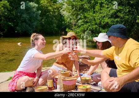 Pique-nique au bord du Doubs et du Canal Charles Quint à Dole (nord-est de la France) : groupe d'amis déjeunant au bord de l'eau en été, avec casquettes Banque D'Images