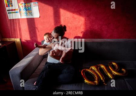 Diversity père se trompant, s'amusant avec son fils sur un canapé gris contre un mur rouge. Un homme célèbre le premier anniversaire d'un petit garçon. Golden Banque D'Images