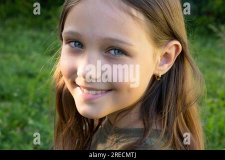 Portrait d'une belle fille brune regardant la caméra et souriant, debout dans la rue sur fond de verdure un jour d'été. Jeune enfant curieux lève les yeux et baissa la tête regarde la caméra en gros plan à l'extérieur. Visage yeux contemplatifs enfant. Banque D'Images