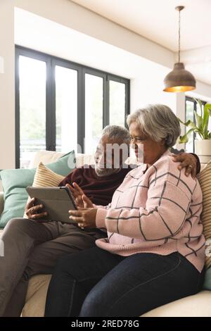 Heureux couple biracial senior assis sur le canapé et ayant un appel vidéo tablette à la maison Banque D'Images