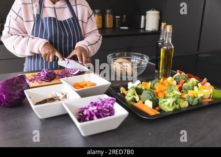 Femme biracial senior portant un tablier coupant le chou sur la table avec des légumes dans la cuisine à la maison Banque D'Images