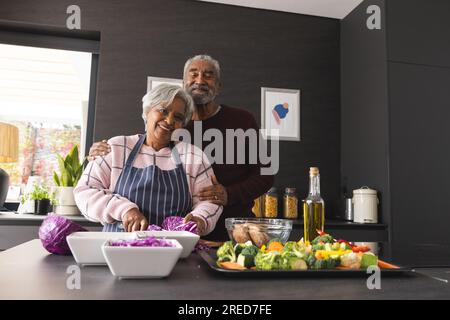 Portrait d'heureux couple biracial senior préparant des légumes et embrassant dans la cuisine à la maison Banque D'Images
